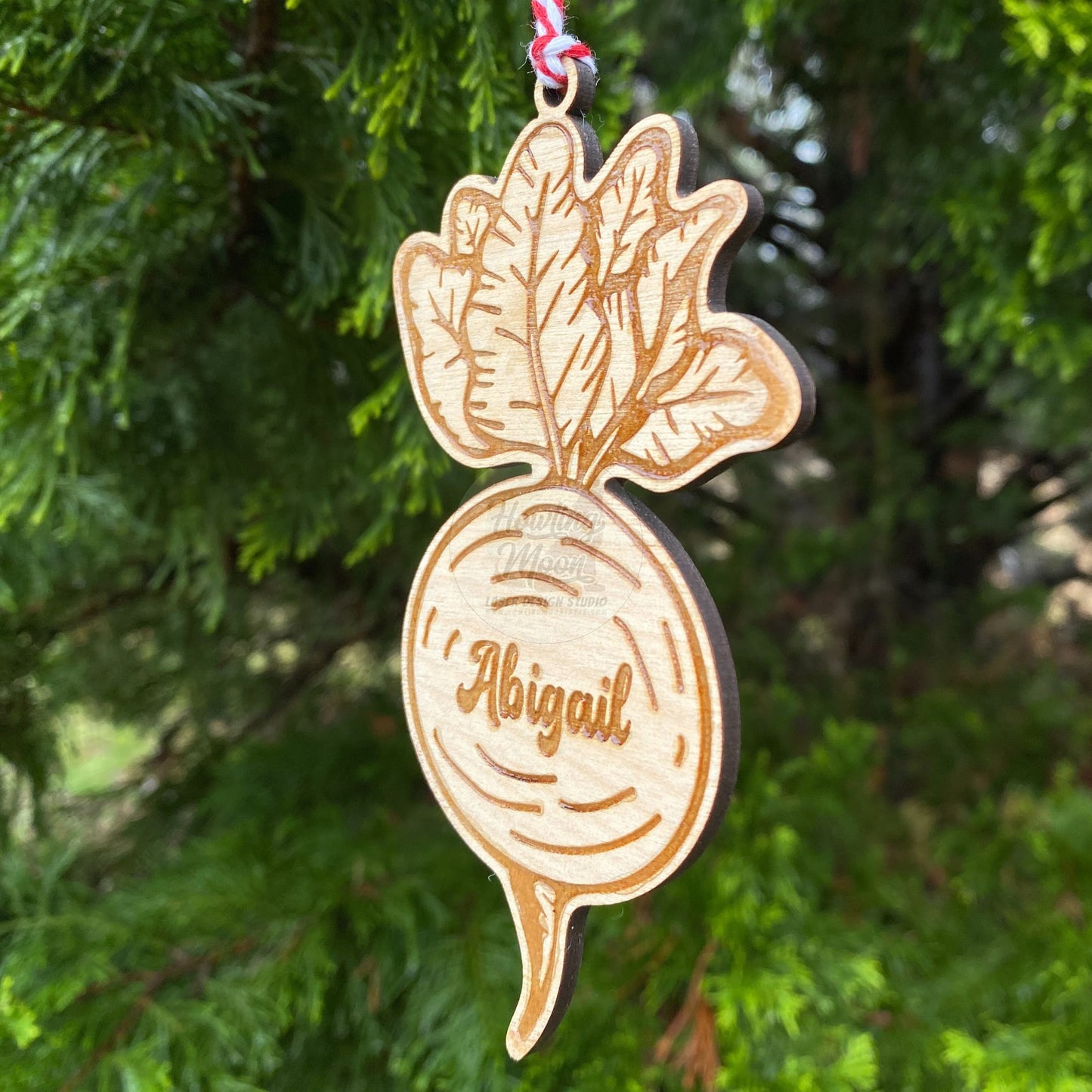 Side view of personalized garden beet ornament with red & white twine ribbon hanging from a coniferous tree - made by Howling Moon Laser Design in Virginia USA